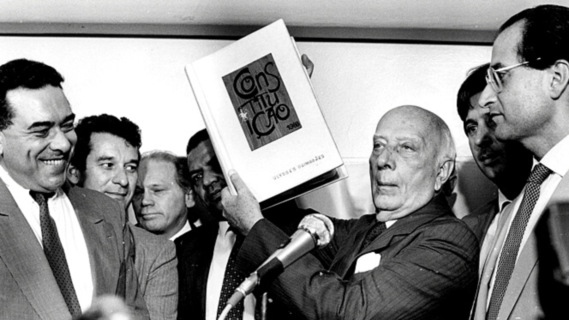 Ulysses Guimarães segurando a Constituição Federal de 1988. Momento importante para democracia brasileira, tal documento garante cobrança progressiva de imposto de renda. Ulysses Guimarães segurando a Constituição Federal de 1988, apresentando documento que garante a cobrança progressiva do Imposto sobre a Renda.