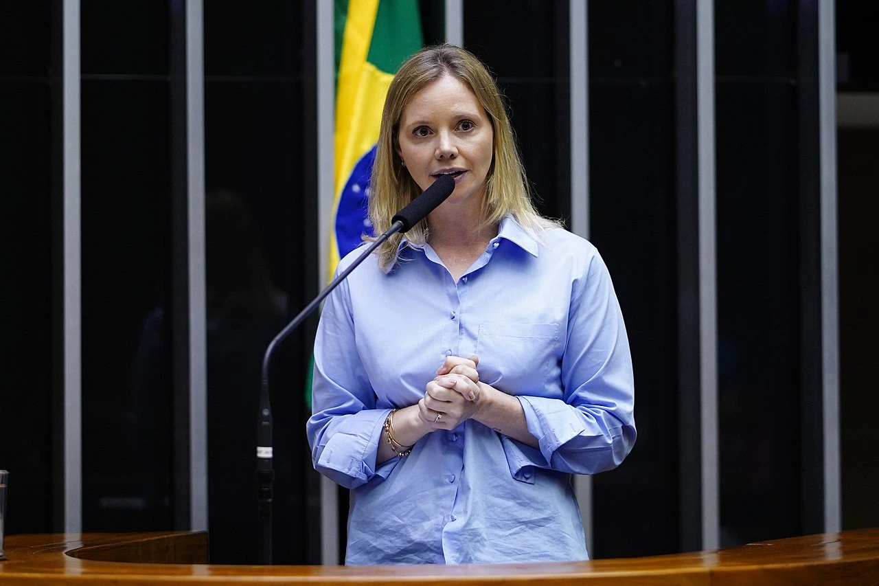 Deputada Dani Cunha (UNIAO - RJ) no plenário da Câmara dos Deputados, onde apresentou o PL 2720/2023 para criminalizar a discriminação contra pessoas politicamente expostas. Usa camisa social azul, em fundo preto com bandeira do Brasil.