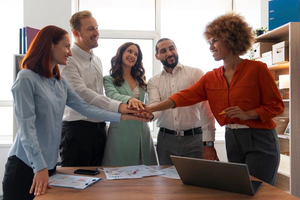 A imagem retrata um grupo de pessoas sorridentes, composto por três mulheres e dois homens, reunidos em local de trabalho acolhedor e promotor de bem-estar, que transmite a sensação de união, parceria e harmonia.