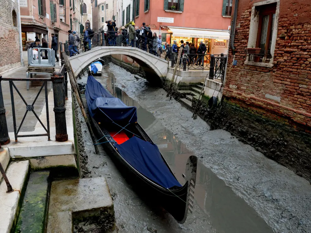 A imagem apresenta um canal da cidade de Veneza, na Itália. Ele está seco e tem um barco no leito do canal. ao fundo pessoas atravessam uma pequena ponte a pé e olham para o canal vazio. 