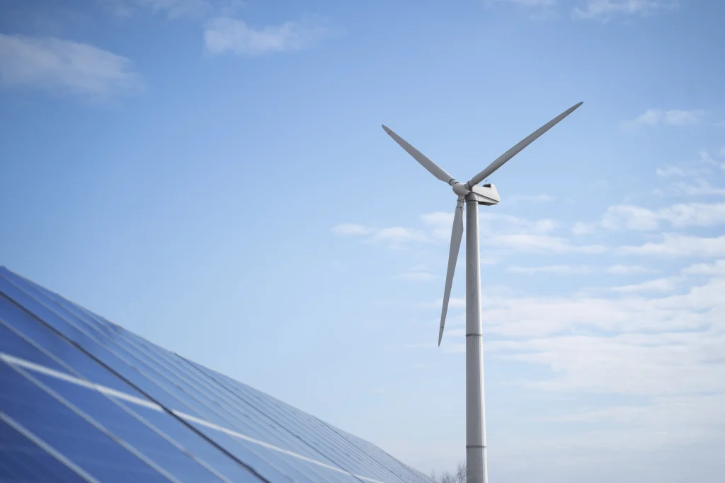 Foto com fundo do céu, à esquerda uma placa de produção de energia solar e à direita produção de energia eólica, evidenciando a diversidade de produção da energia limpa 