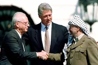 A imagem mostra um aperto de mãos entre Yitzhak Rabin, primeiro ministro de Israel e Yasser Arafat, líder da OLP. Os dois ocupam extremidades opostas da imagem e no centro, entre os doi, está Bill Cliton. 