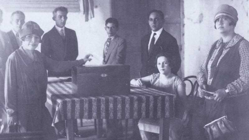 Foto antiga de mulheres e homens em volta de uma mesa com uma urna ao centro, votando.