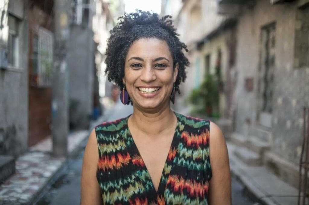 Foto de Marielle Franco, mulher negra de cabelos crespos curto, usando um vestido e sorrindo na frente de uma favela.