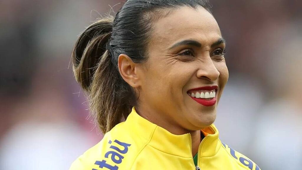 Marta com casaco da seleção brasileira sorrindo com batom vermelho.