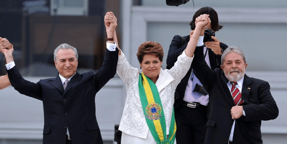 A imagem mostra uma foto de Michel Temer, Dilma Rousseff e Luiz Inácio Lula da Silva. Os três estão lado a lado, de mãos dadas e elevadas acima da cabeça.