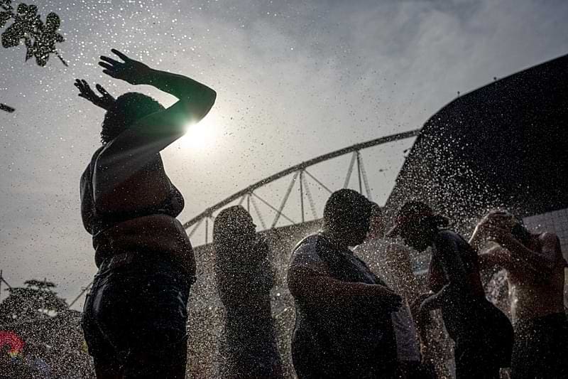 Pessoas na frente de estádio sendo molhadas por uma mangueira, ao fundo o sol apresentando uma situação de calor.