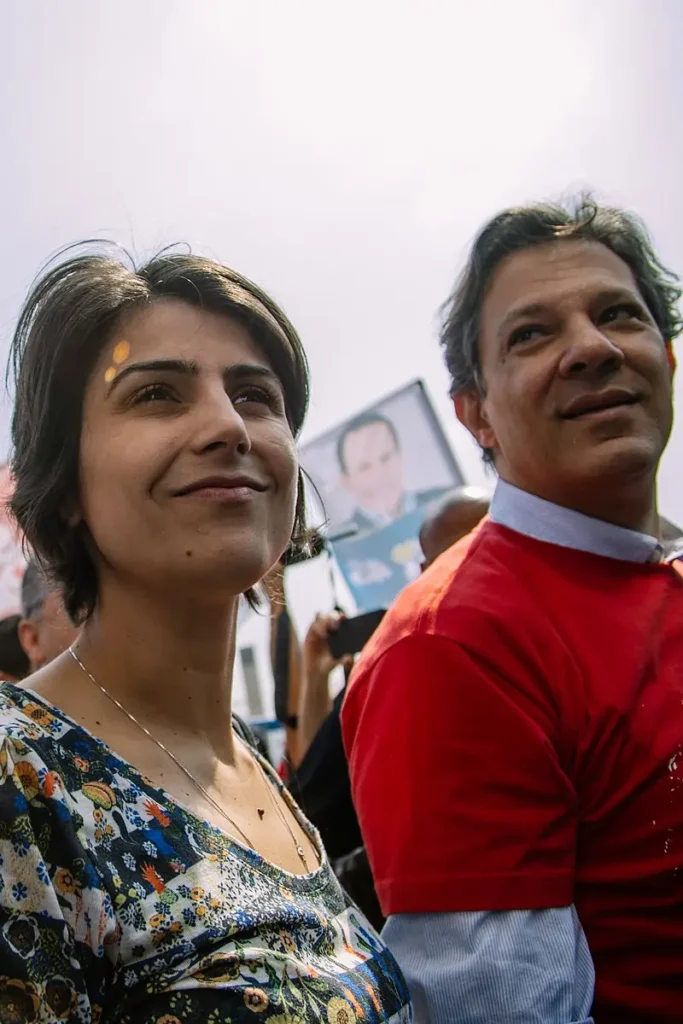 Fotografia de Manuela d’Ávila  e Fernando Haddad em um evento de campanha eleitoral ao ar livre. d’Ávila, em primeiro plano, usa uma blusa floral; Haddad, ao seu lado, veste camisa vermelha.