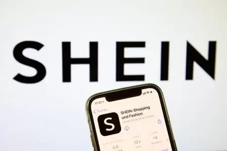 Loja da shein Logo da loja Shein ao fundo e a frente está um celular que mostra o aplicativo da loja.