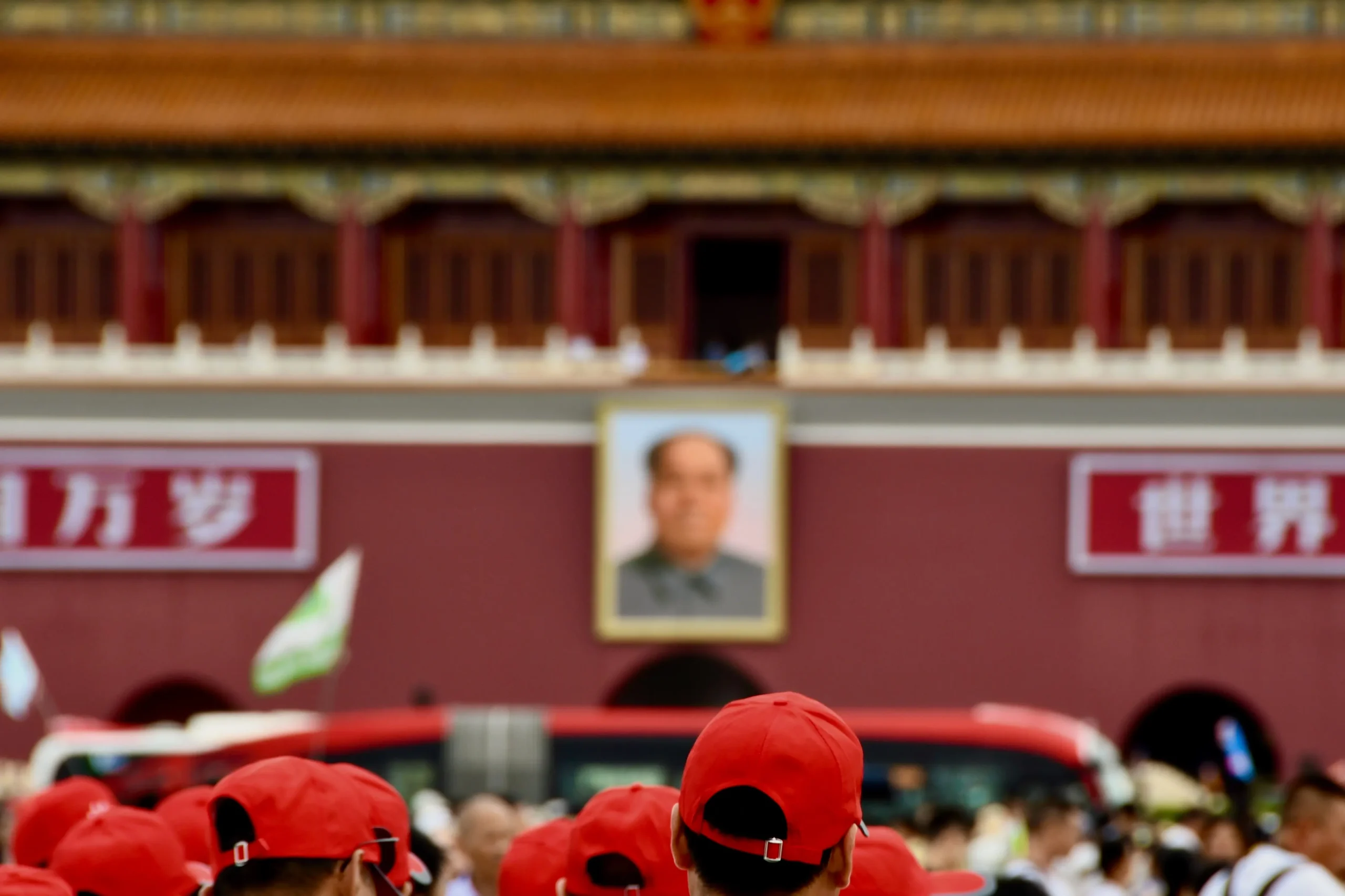 a imagem apresenta, em um plano principal, um prédio com uma imagem de Mao Tsé-Tung. Abaixo, percebe-se uma multidão em deslocamento. No primeiro plano da imagem, as  pessoas usam bonés vermelhos.