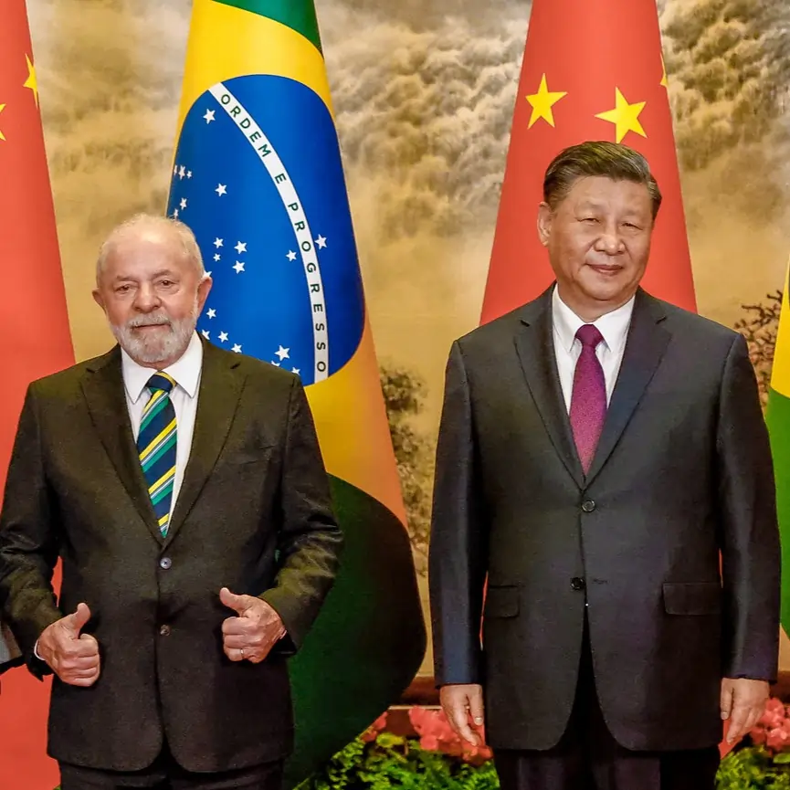 A imagem apresenta o presidente do Brasil, Lula ao lado do presidente da China, Xi Jinping. Os dois estão lado a lado e na frente das bandeiras de seus respectivos países. Lula aparece com os polegares levantados 