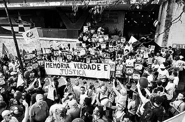 A imagem mostra uma multidão de pessoas que seguram faixas e cartazes. Um deles diz "memória, verdade e justiça".