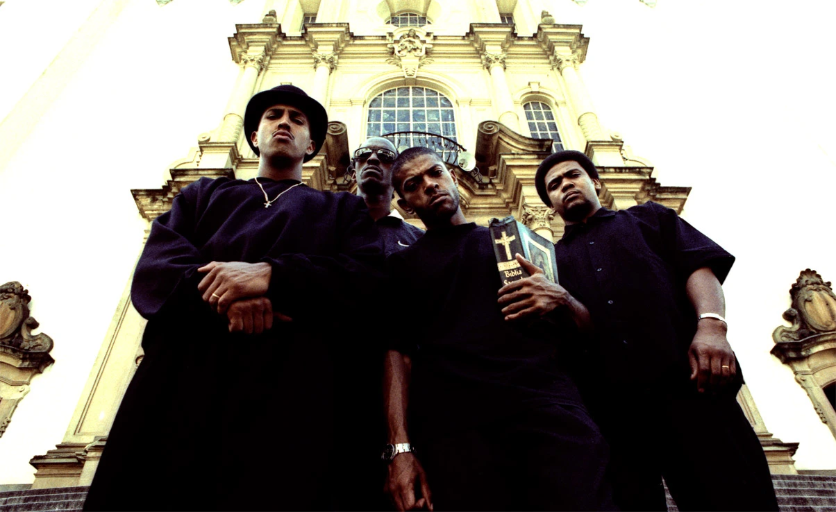 Grupo Racionais MC's Fotografia de quatro homens negros em frente a uma igreja.