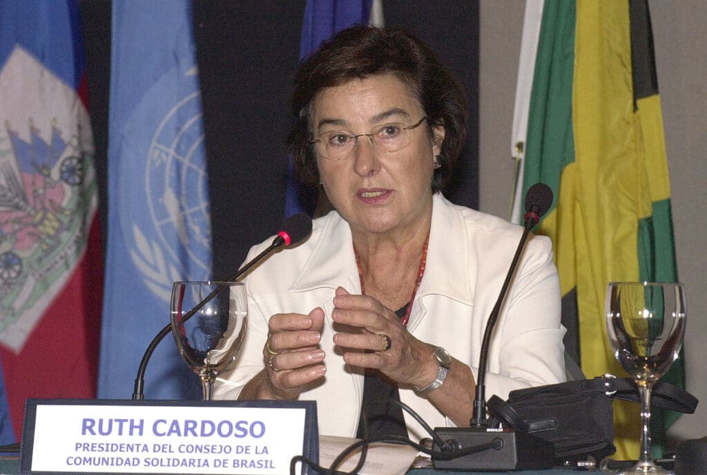 A imagem mostra Ruth Cardoso, ex primeira-dama do Brasil em um evento do projeto Comunidade Solidária. Ela está sentada diante de uma mesa e de um microfone, onde ela fala. Ao fundo bandeiras aparecem, uma delas é da ONU. 